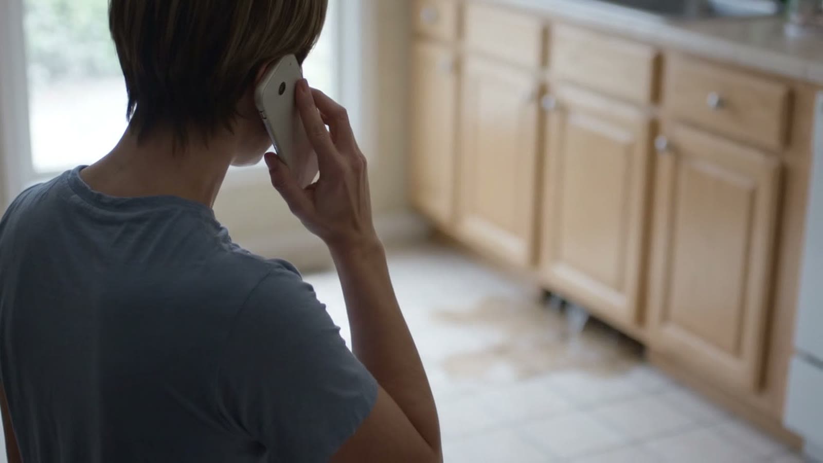 A woman standing in her kitchen holding a phone to her ear, waiting.