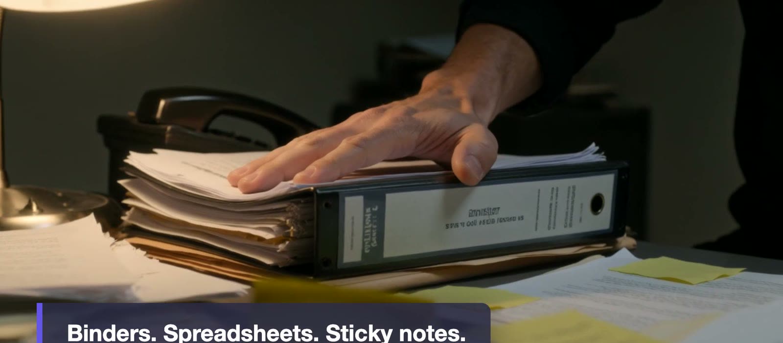 A pair of hands resting on a thick stack of binders and loose paperwork on a desk.